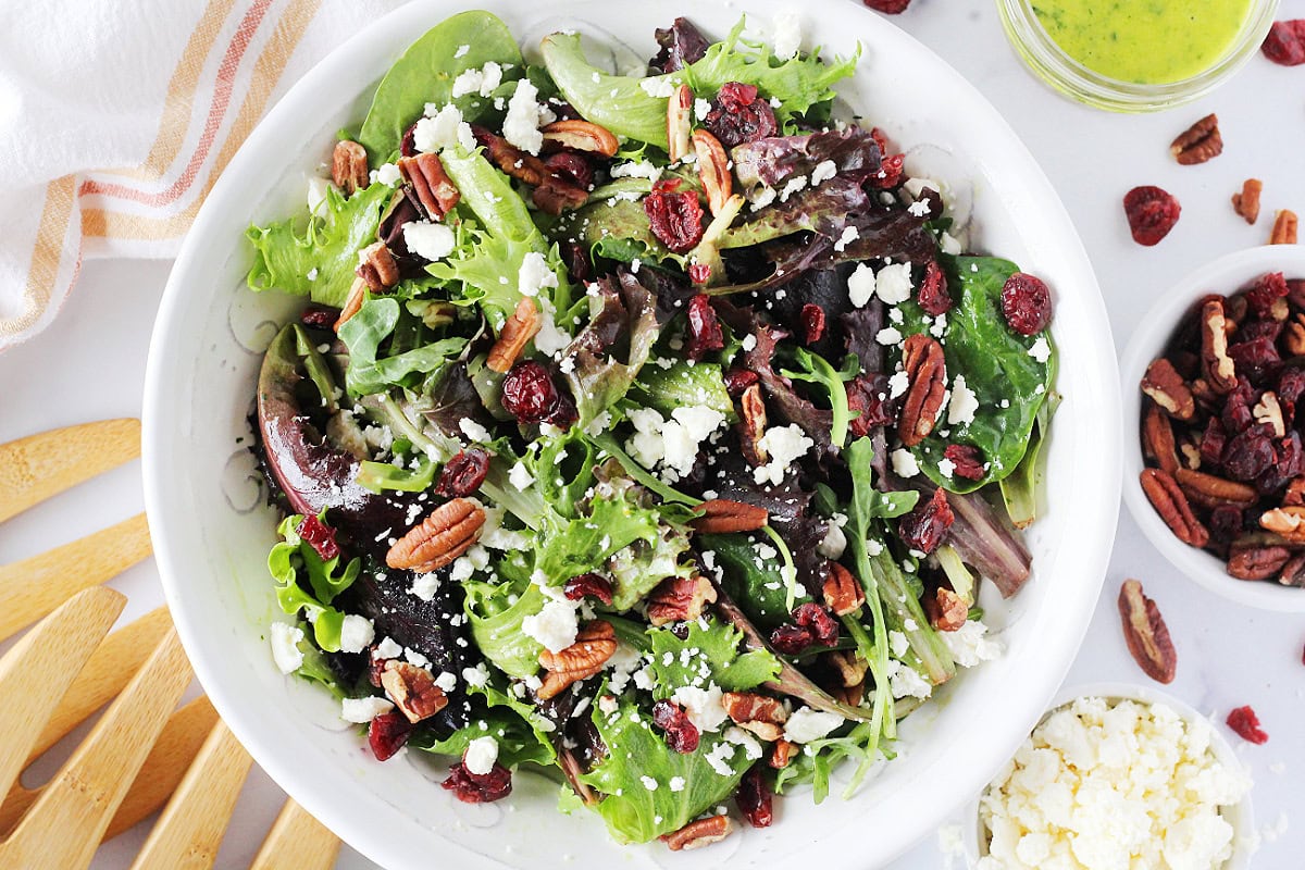 Mixed greens in a bowl topped with crumbled feta cheese, dried cranberries, and pecans.