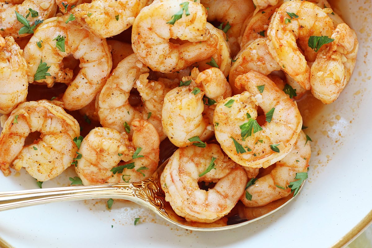 A gold spoon in a white bowl of saucy oven baked shrimp.