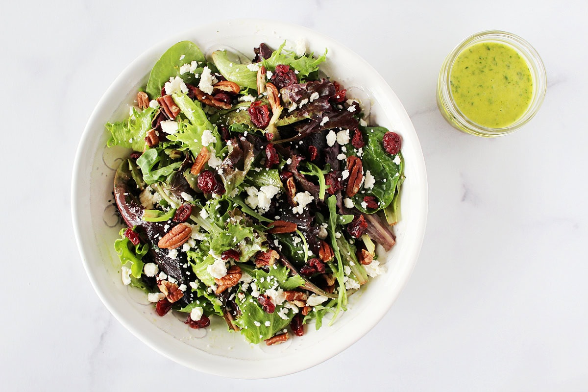 Citrus dressing beside mixed green salad topped with feta, pecans, and dried cranberries.