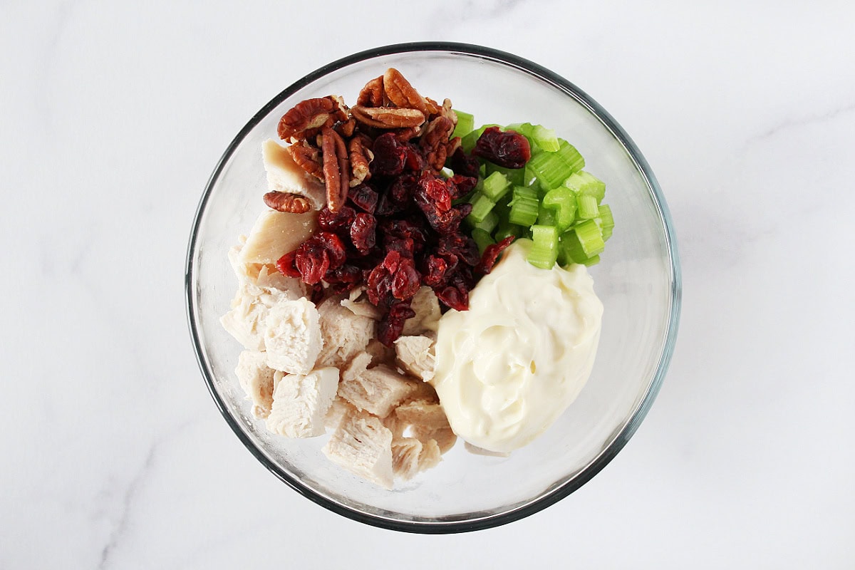 Diced chicken breast, mayo, celery, pecans, and dried cranberries in a glass bowl.
