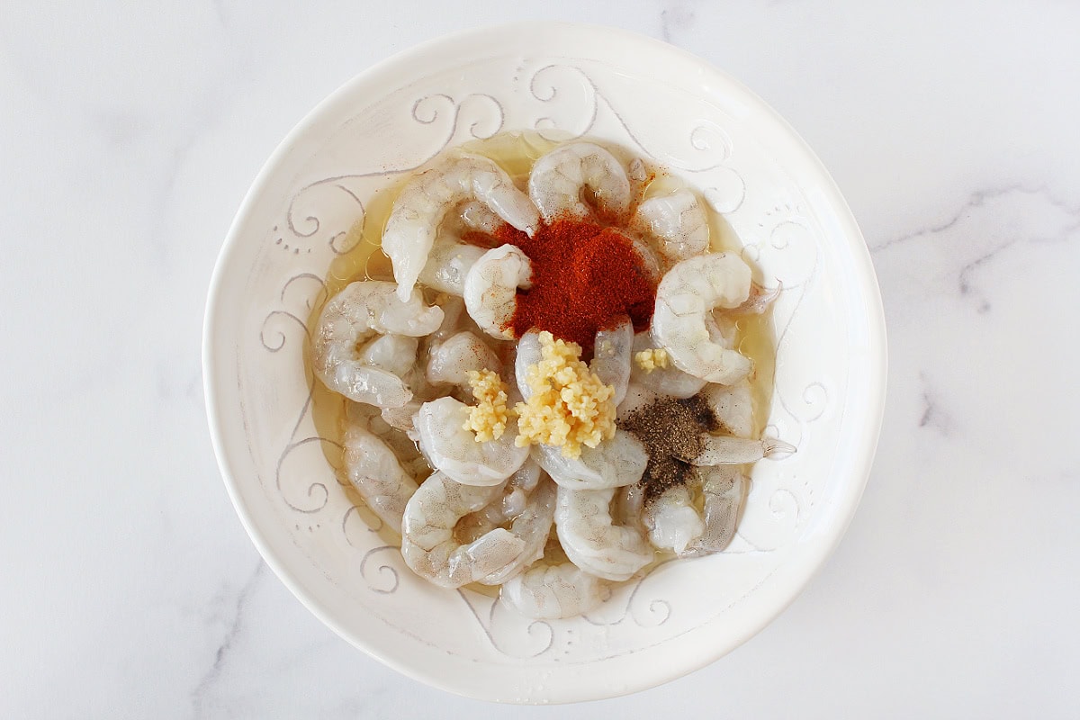 Raw shrimp, lemon juice, minced garlic, paprika, and black pepper in a bowl.