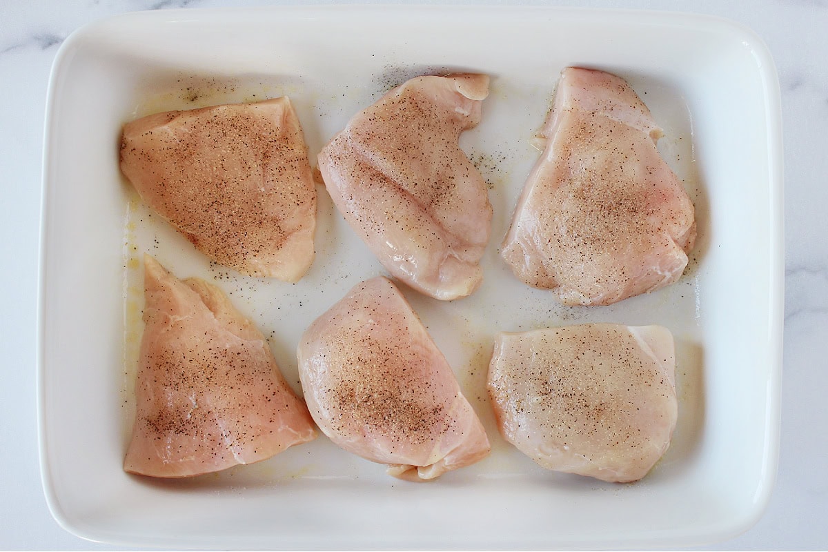 Six raw chicken breasts topped with salt and black pepper in a white baking dish.