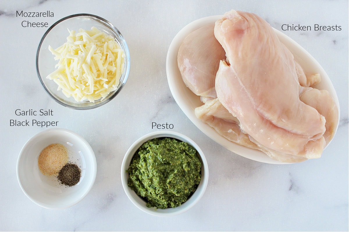 Labeled ingredients for baked pesto chicken breast on a white marble countertop.