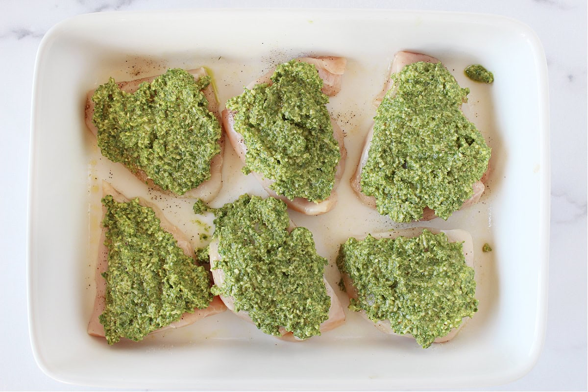 Homemade walnut pesto on top of six raw chicken breasts in a baking dish.
