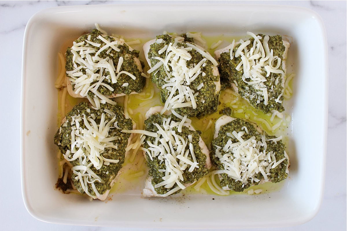 Shredded mozzarella on top of baked pesto chicken in a baking dish.